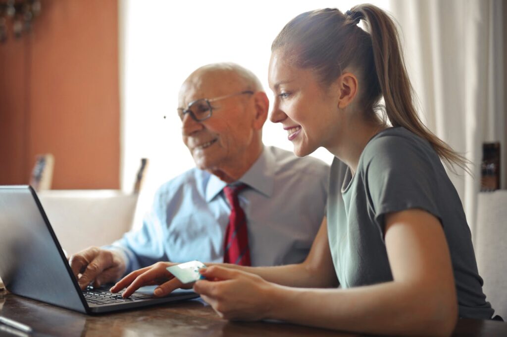 pexels-photo-3823488 Young woman in casual clothes helping senior man in formal shirt with paying credit card in Internet using laptop while sitting at table